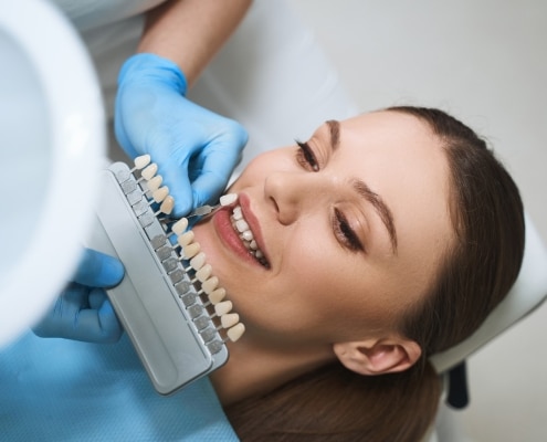 cheerful-young-lady-is-lying-dental-chair-choosing-veeners-colour-with-doctor-help